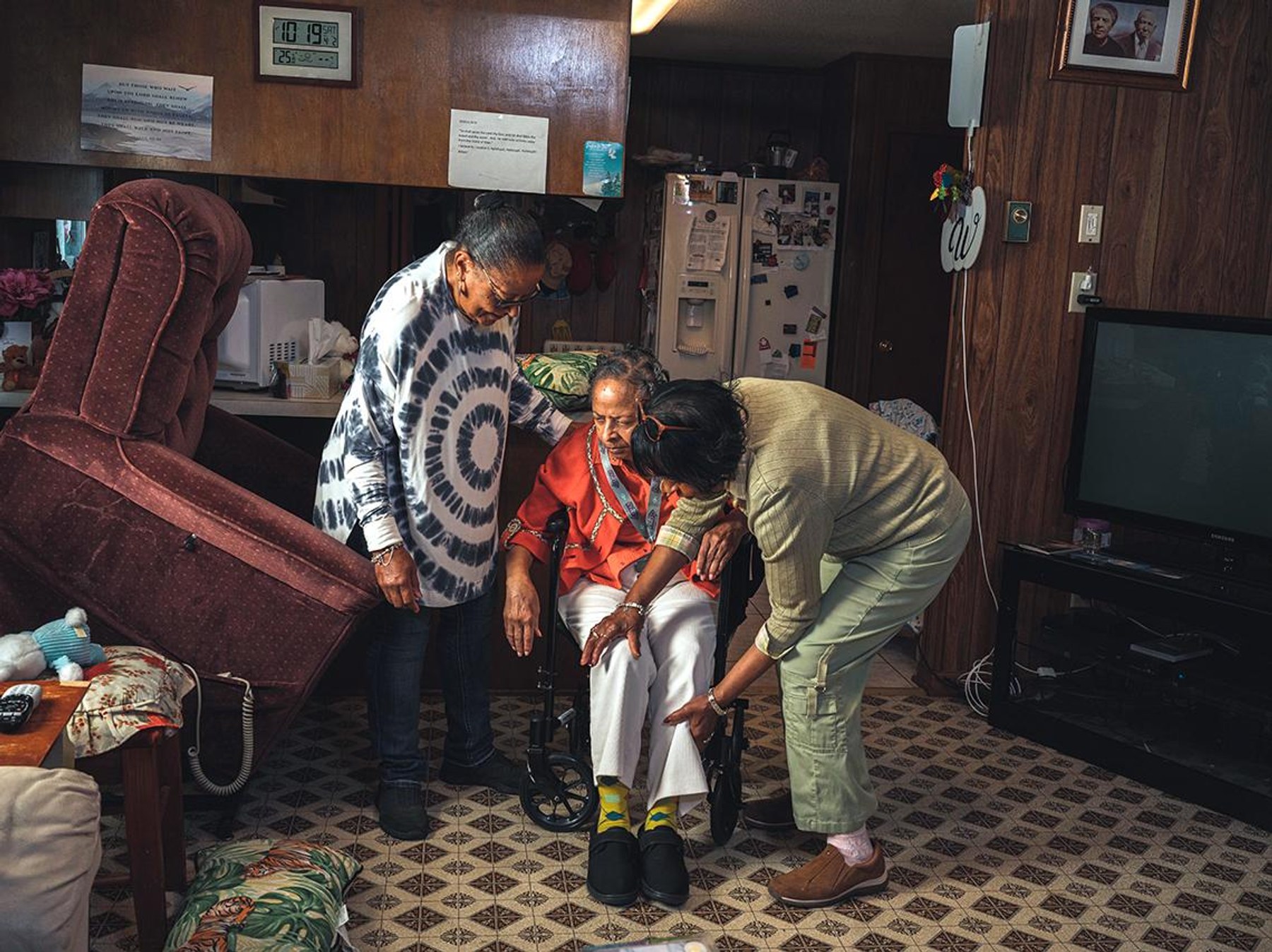 Two family caregivers help an older woman stand up with her walker at home.
